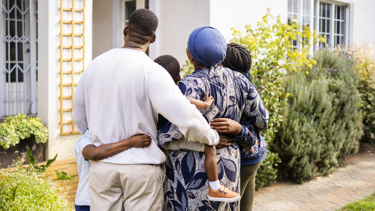 A family of five stands in front of a home with their arms wrapped around each other.  
