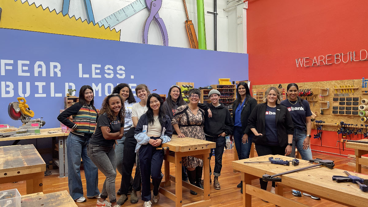 U.S. Bank employees and Girls Garage staff stand in the Girls Garage workshop surrounded by tools and woodworking stations. Background mural reads: Fear Less. Do More. 