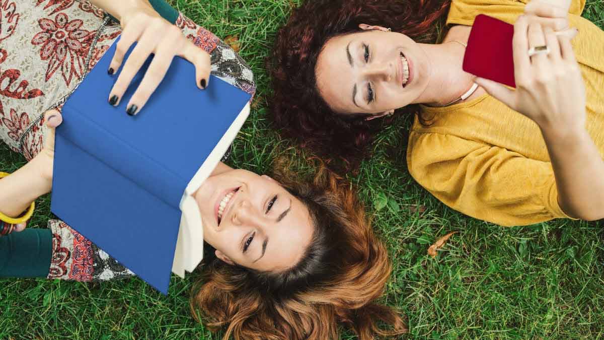 Two women lying in the grass reading a phone and a book, respectively