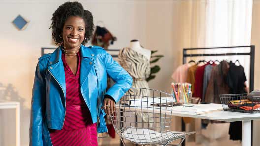  Young Black female clothing designer poses in her studio, smiling. She wears a vibrant blue jacket.