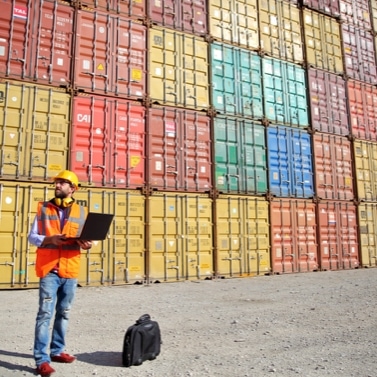 Person holding laptop standing in front of stack of shipping containers