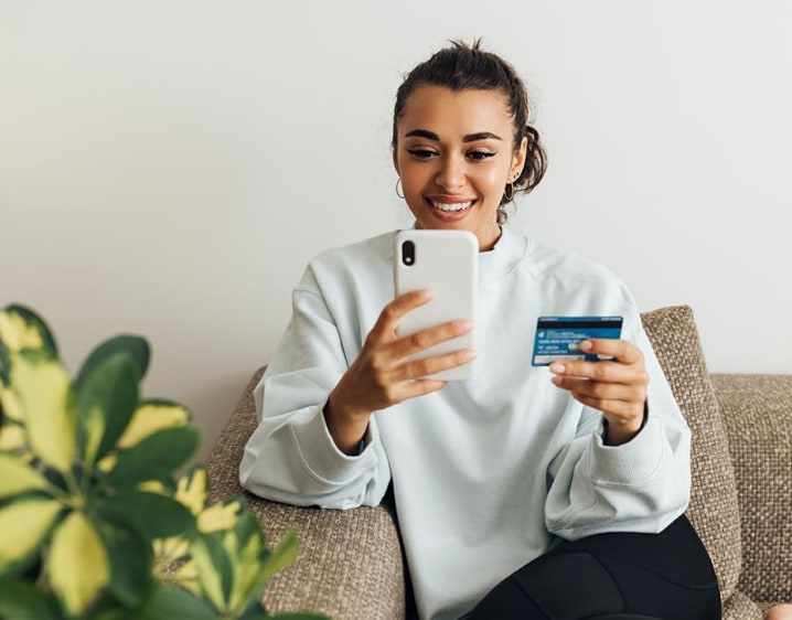 Young woman sitting on her sofa with a credit card in one hand and her phone in the other. She is using her phone to pay a bill.