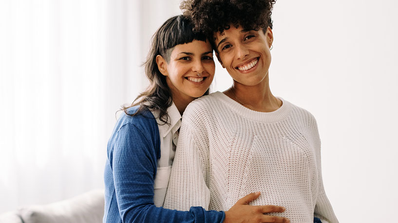 A pregnant female couple smiles at the camera.
