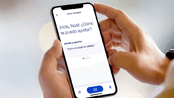 A cell phone screen showing the U.S. Bank Mobile App faces the camera (only the hands of the person holding it are shown). The screen says: ‘Hola, Noel! Como te puedo ayudar?’ and features a speaking prompt below, also in Spanish.