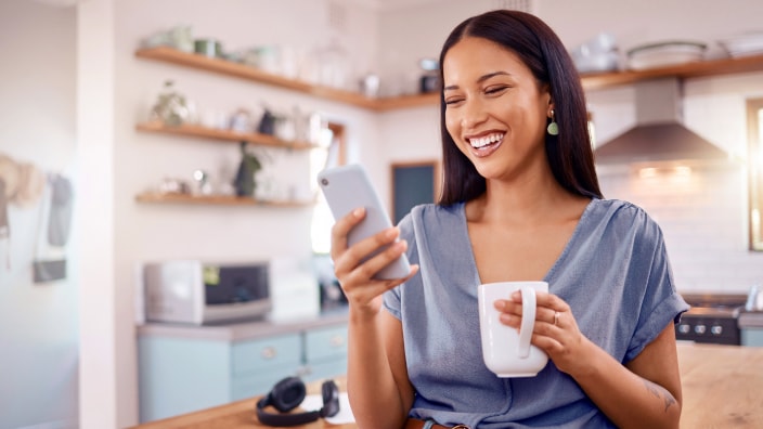A young Hispanic woman explores Financial IQ on her phone while holding a mug in her kitchen